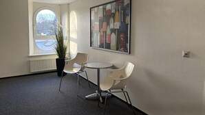 Ein Zimmer mit rundem Tisch und zwei Stühlen, einem großen abstrakten Gemälde an der Wand, einer Topfpflanze und einem Fenster mit Blick auf ein Auto draußen.
