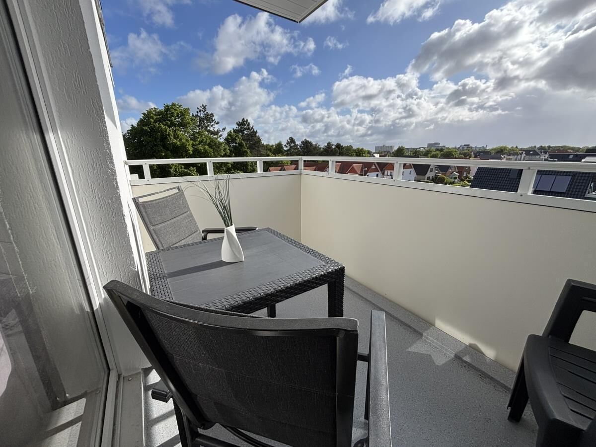 A balcony with a table and chairs, a white railing, and a view of buildings and trees.
