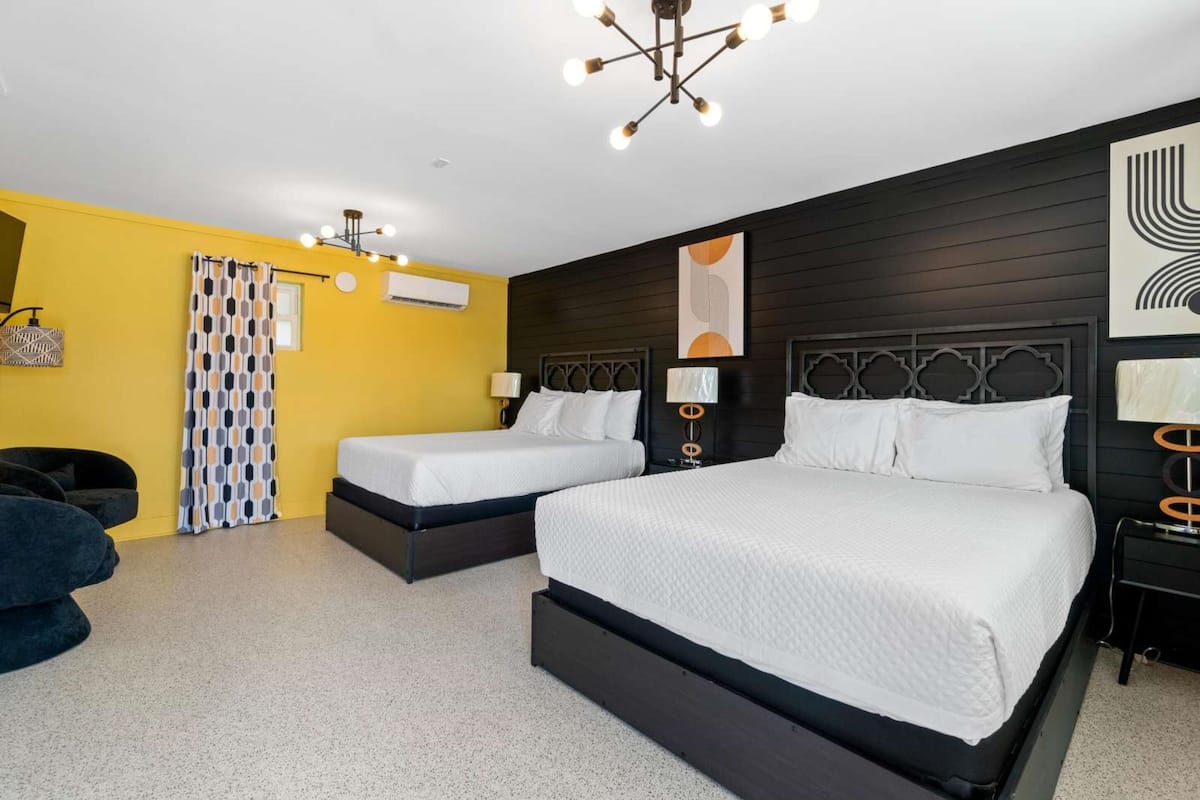 Een hotelkamer met twee bedden, een donker houten hoofdbord, een geel accentmuur en een moderne kroonluchter.