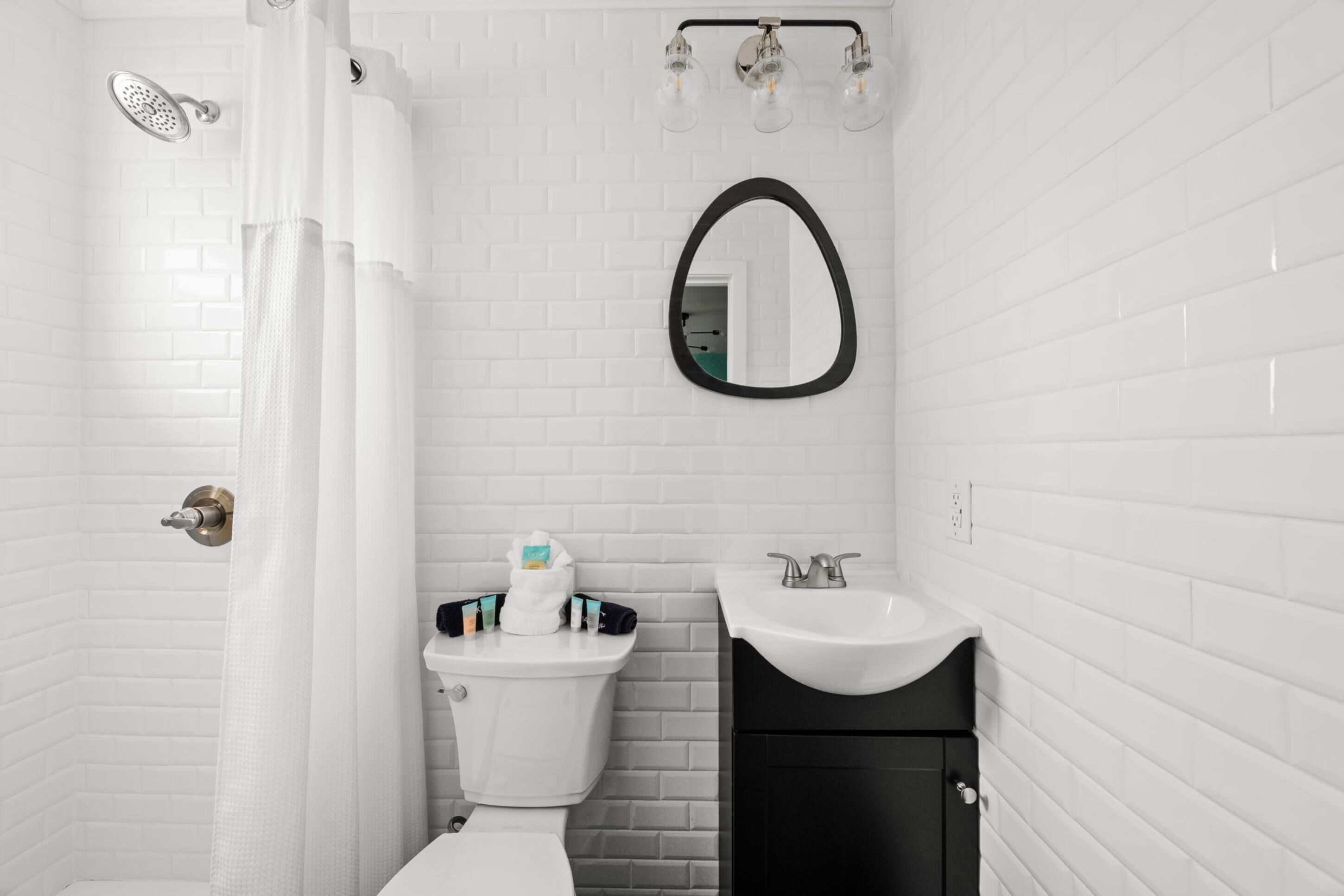 Casa de banho com azulejos brancos tipo metrô, toucador preto, sanita branca, chuveiro com porta de vidro e espelho com moldura preta.