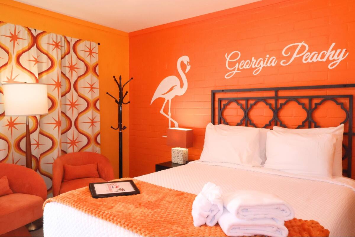 Hotelkamer met een bed, nachtkastje, lamp, stoel en een muur met een flamingo en de tekst 'Georgia Peachy'.