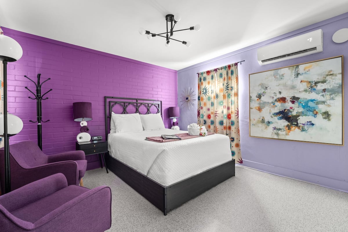 Een slaapkamer met een paarse bakstenen muur, een bed met witte beddengoed, een moderne kroonluchter, een nachtkastje met een lamp, een paarse fauteuil, en een groot abstract schilderij.