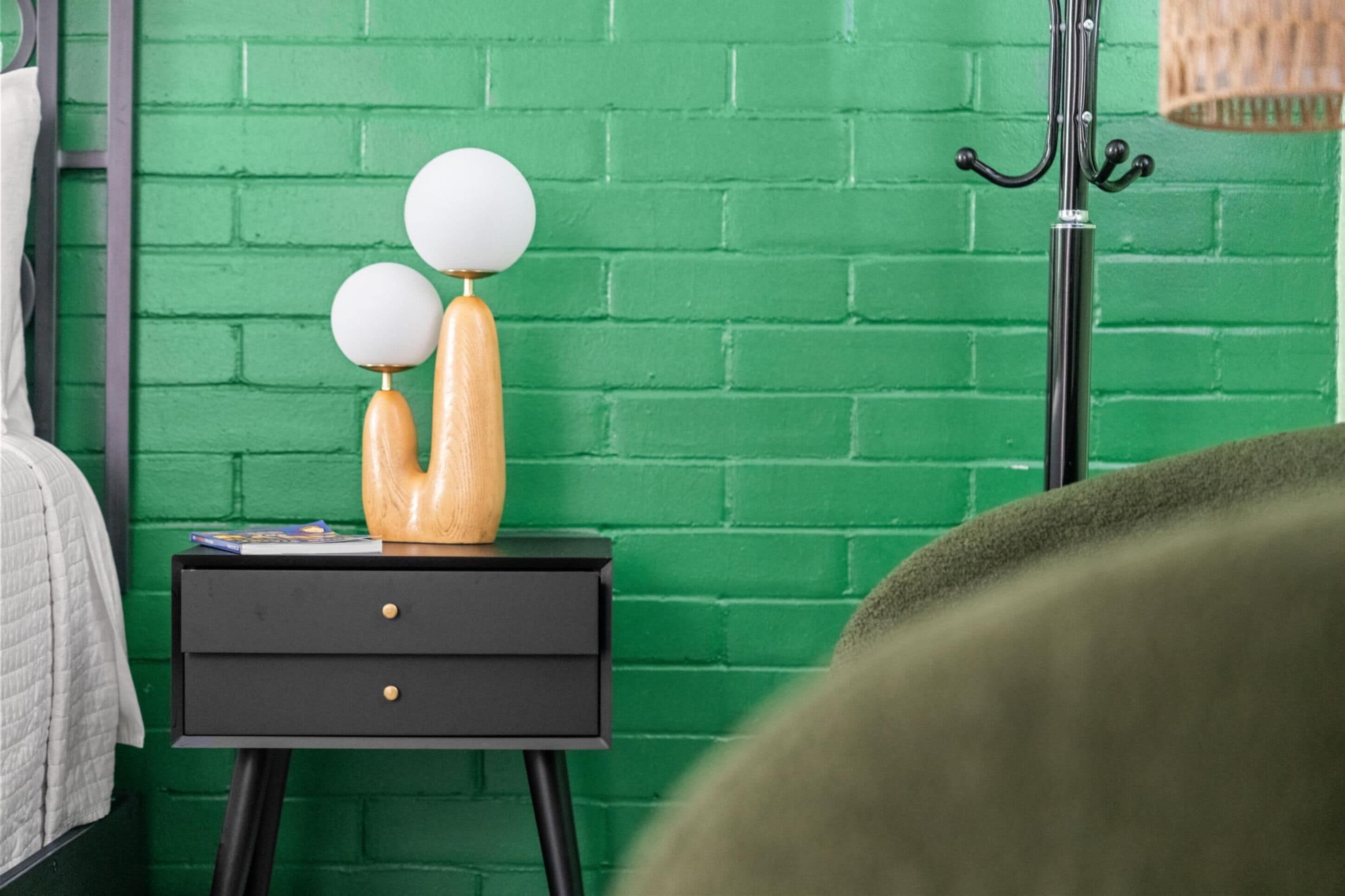 Un mur en briques vertes, une table de chevet noire sur laquelle reposent deux objets sphériques, et un porte-manteau en métal.