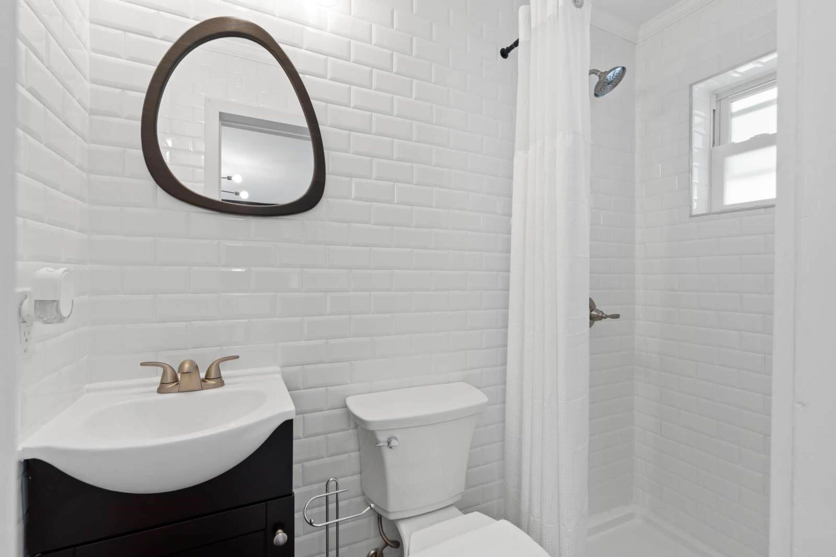 Un baño con azulejos blancos tipo metro, un espejo redondo, un lavabo con grifo dorado y un inodoro.