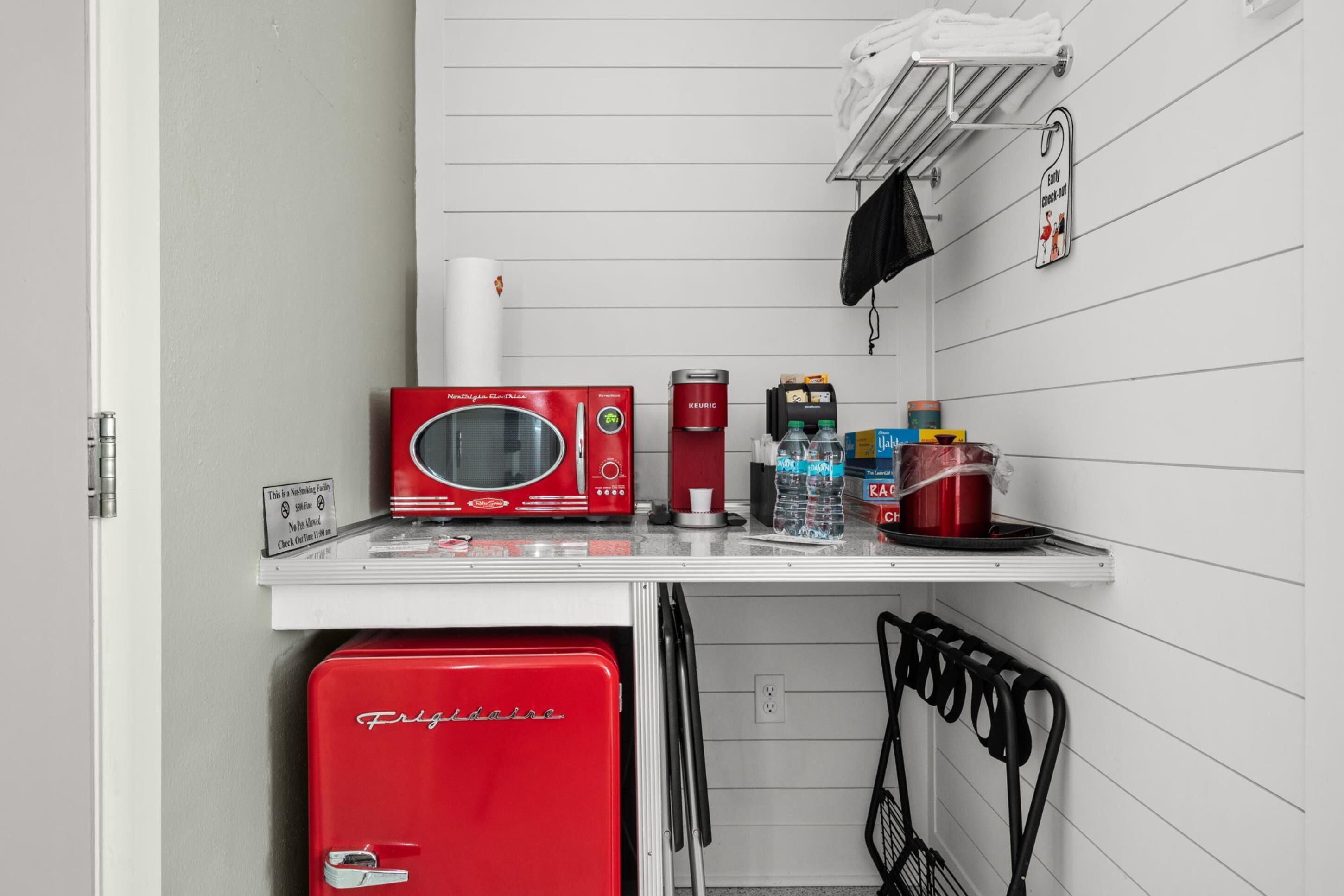 Una kitchenette con un refrigerador Frigidaire rojo, un microondas rojo, una cafetera Keurig, agua embotellada y una tetera.
