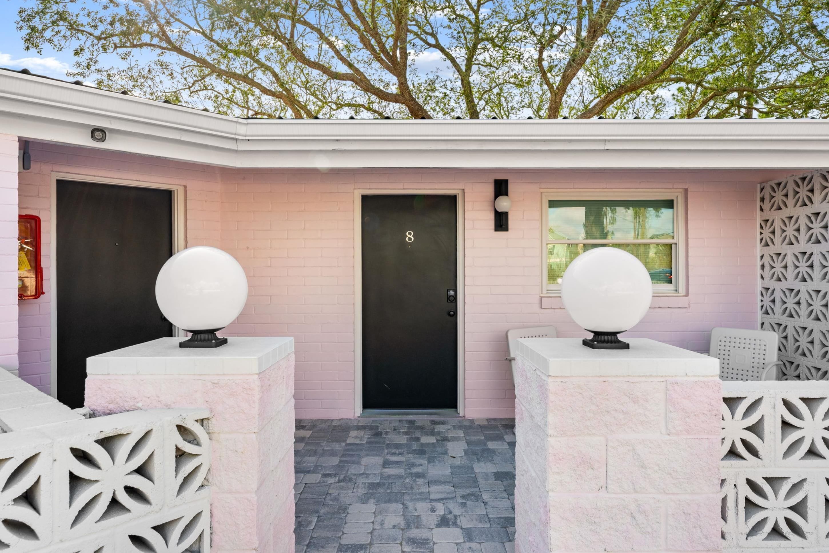 A pink brick building with a black door marked with the number 8, a window, and decorative spherical objects on pillars.