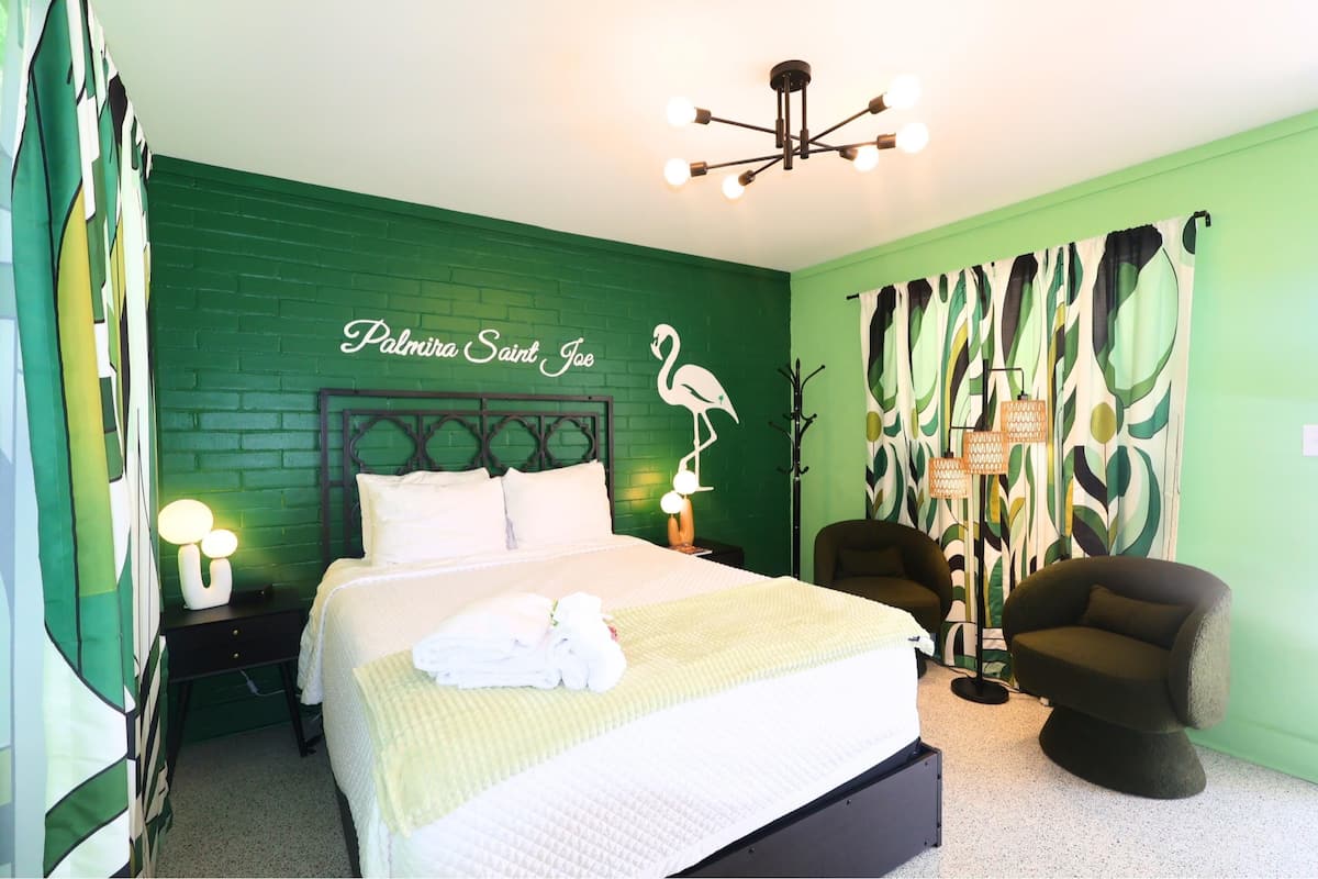 Een hotelkamer met een groene bakstenen muur, een bed met wit beddengoed, een nachtkastje met een lamp, een stoel en een muurschildering.