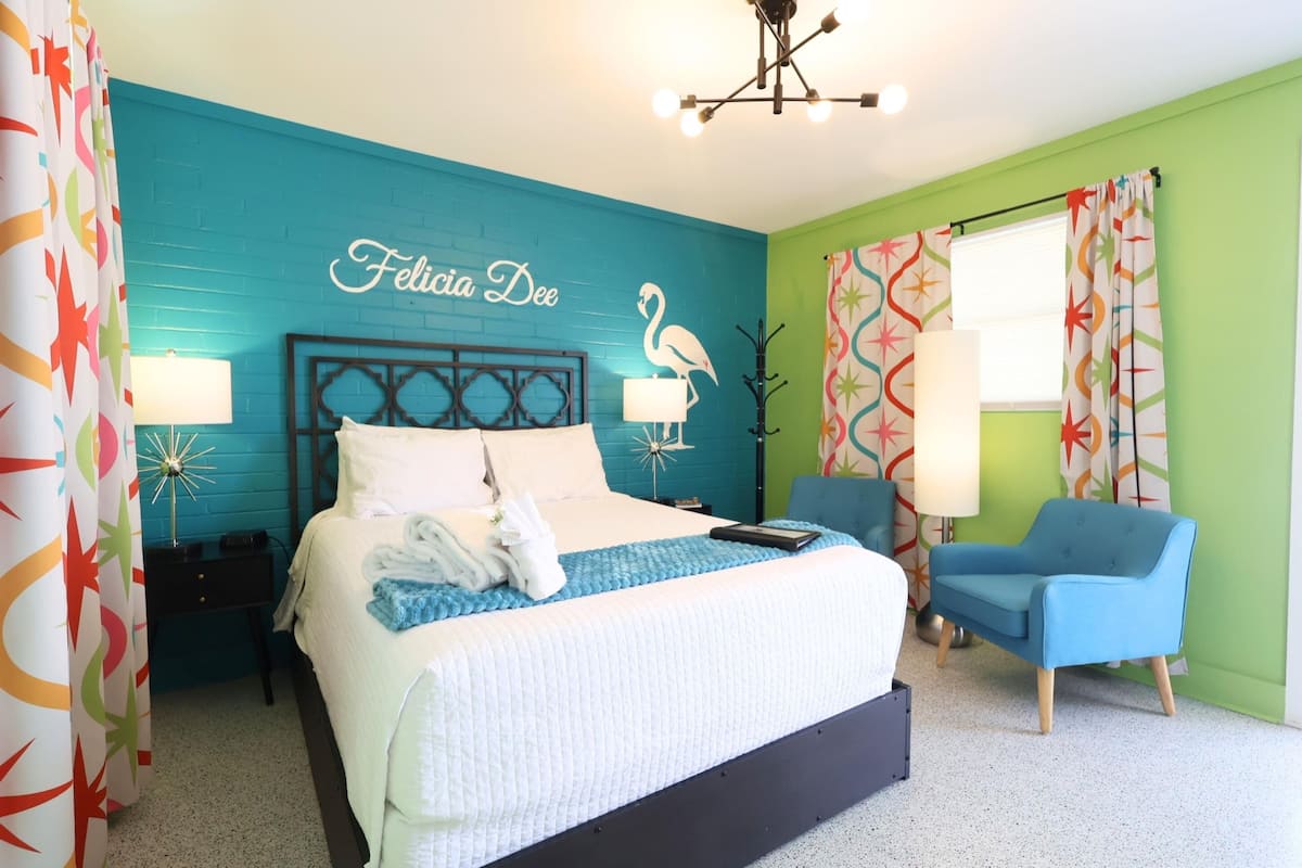 Een slaapkamer met een bed, een stoel, een nachtkastje, een lamp en een wand met een naam en flamingo decoratie.