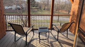 Eine überdachte Terrasse mit zwei Stühlen und einem kleinen Tisch, mit Blick auf einen umzäunten Garten mit Bäumen und einem weit entfernten Haus.