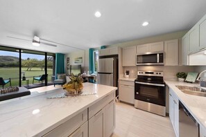 A modern kitchen with a large island, stainless steel appliances, and a view of a green landscape. - FALL into October Savings! Turtle Bay’s Kuilima Estates West #104 (Kahuku)