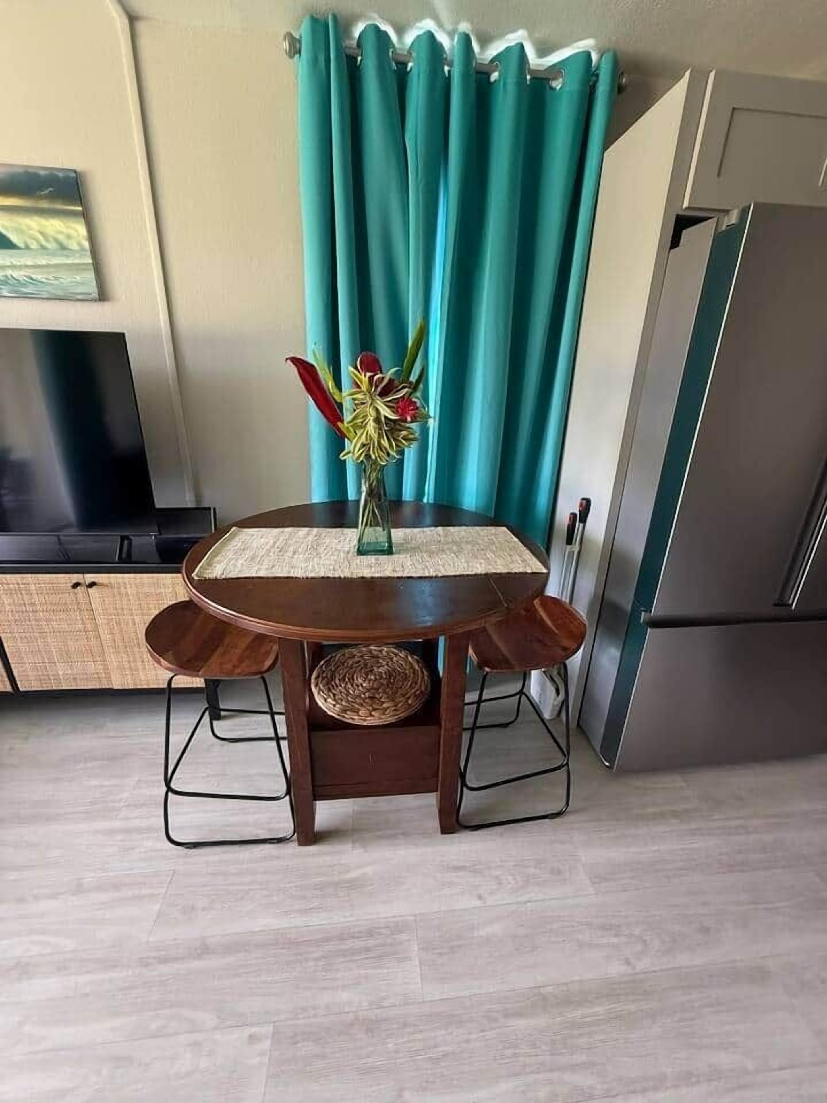 Un comedor con una mesa redonda, dos sillas, un florero con flores, un mantel tejido y una cortina color turquesa al fondo.