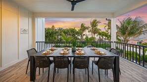 Un coin repas avec une table dressée pour quatre personnes, donnant sur une plage bordée de palmiers et offrant une vue sur le coucher du soleil.