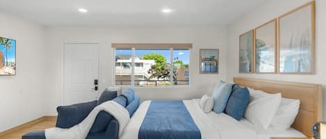 A modern hotel room with a large bed, a sofa, a desk, and a window with a view of buildings and trees.
