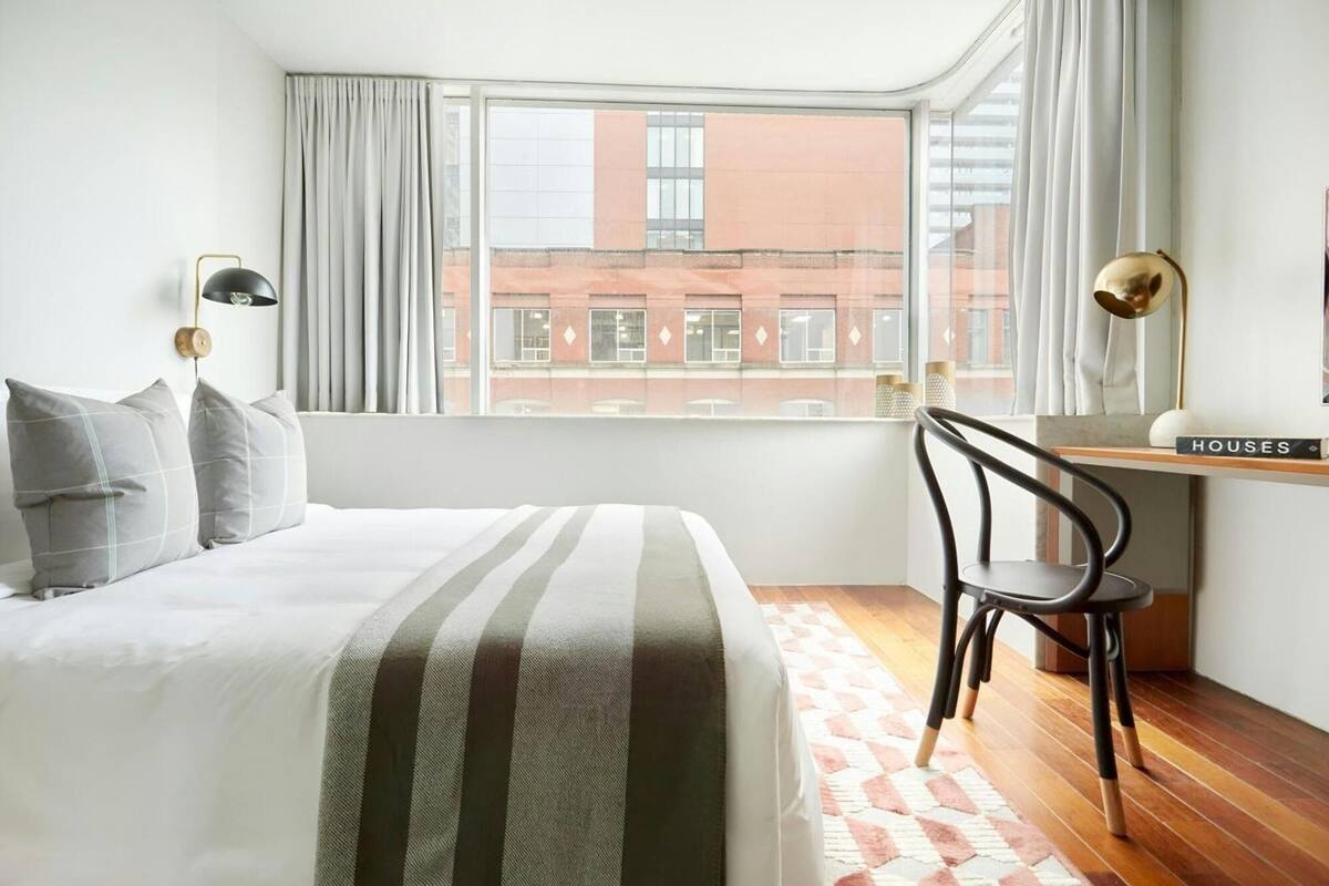 A modern bedroom with a large bed, a desk, and a chair. There is a window with curtains, and a view of buildings outside.