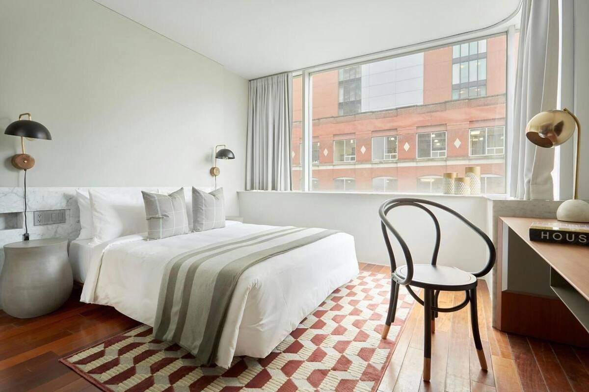 A modern bedroom with a large bed, two bedside lamps, a chair, a desk, and a view of a red brick building through the window.