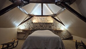 Une chambre confortable située sous les toits, avec un plafond en pente, des poutres apparentes, un mur en pierre et un lit recouvert d’une courtepointe blanche.