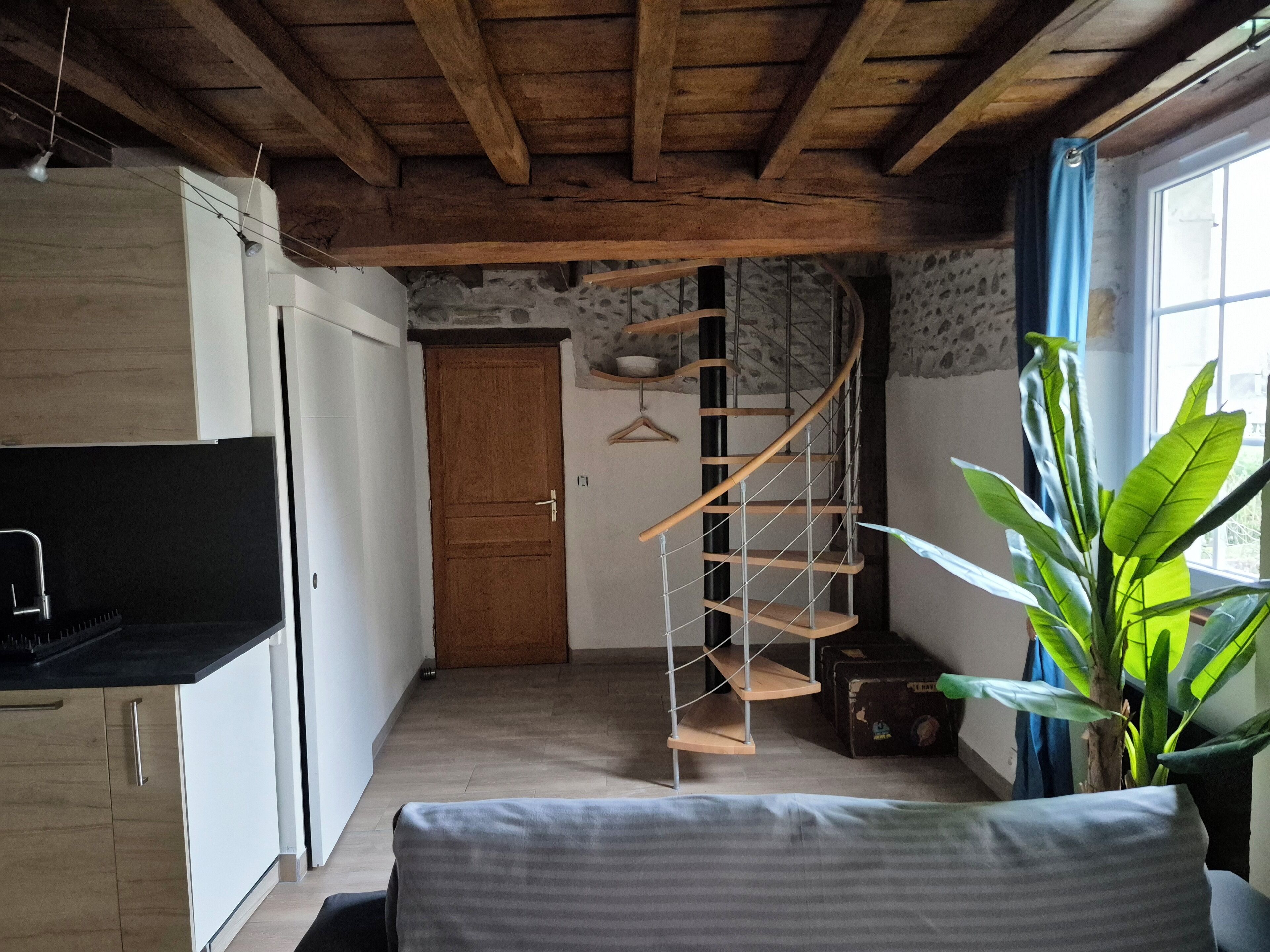 Een compacte kamer met houten plafond, een spiraaltrap, een bed, een potplant en een raam met gordijnen.
