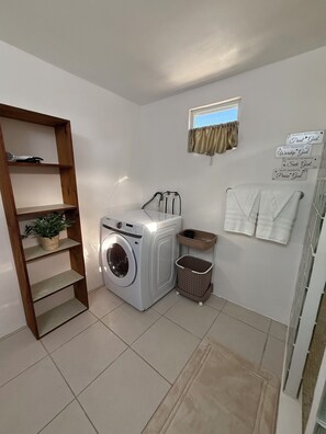 Een wasserette met een wasmachine, een plankje met een plant, een raam met gordijnen, en een muur met motiverende bordjes.