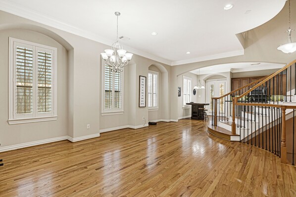 Amplia sala de estar con pisos de madera, ventanas grandes con contraventanas, un candelabro y una escalera con barandilla de madera.