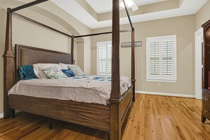 Una cama con dosel de madera con un edredón color beige, dos ventanas con contraventanas blancas y suelo de madera.