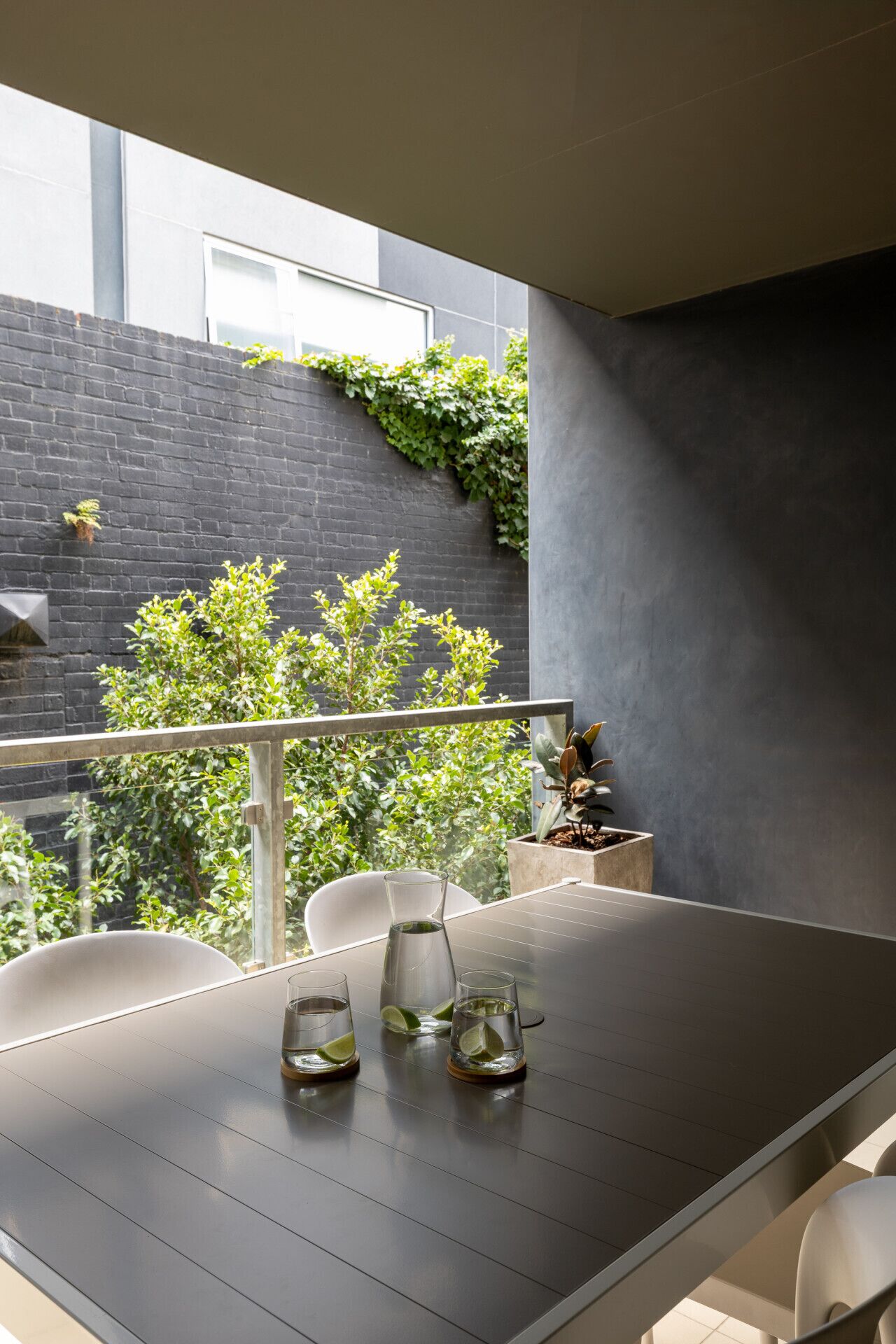 A modern balcony with a dark grey wall, a metal railing, and a table with two glasses of water and lime slices.