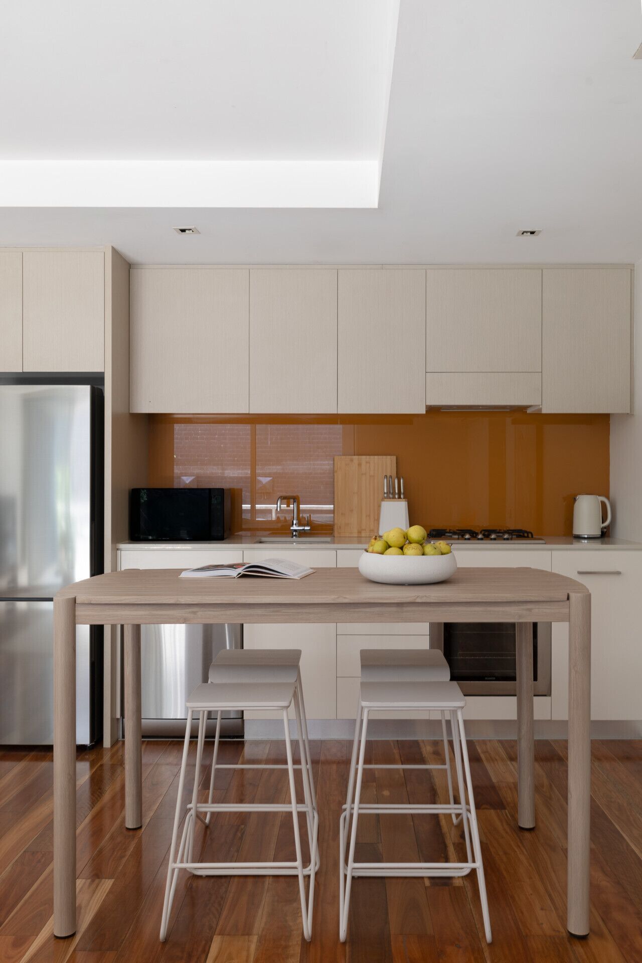 Una cocina moderna con una mesa de madera, dos taburetes blancos, un bol de fruta y una estufa.