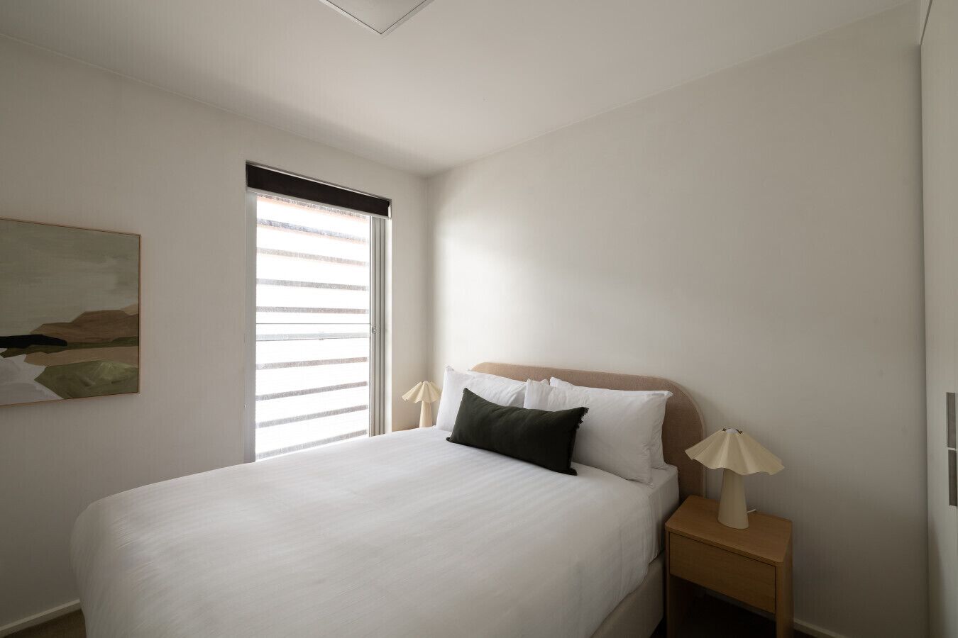 A neatly made bed with white linens and a dark pillow, a bedside lamp, and a painting on the wall.