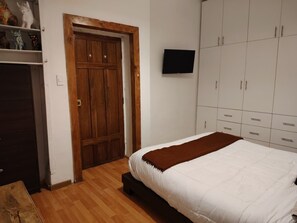 A bedroom with a wooden wardrobe, a bed with a white and brown blanket, a television on the wall, and a wooden floor. - KANTIQ House (Cusco)