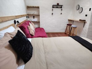 Un dormitorio con cama, cabecera de madera, estante con vasos, una mesita y taburetes.