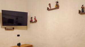 A bedroom with a bed, a TV mounted on the wall, a wooden dresser, and decorative items on shelves.