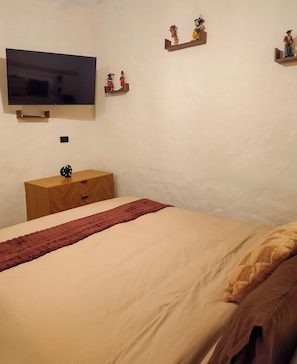Un dormitorio con cama, televisión montada en la pared, cómoda de madera y objetos decorativos en estantes.
