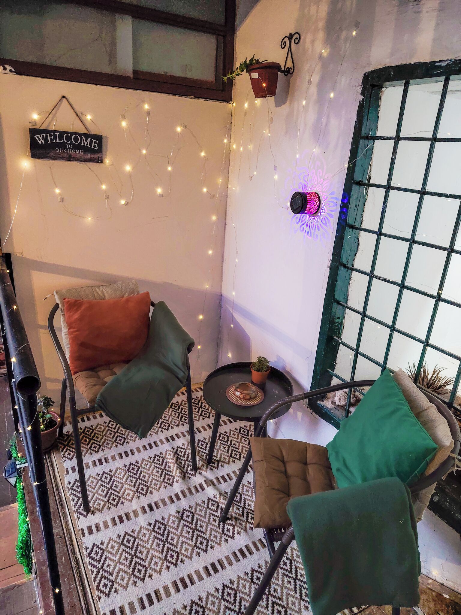 Un coin accueillant avec une pancarte indiquant « BIENVENUE CHEZ NOUS », des guirlandes lumineuses, un tapis à motifs et deux fauteuils avec des coussins.