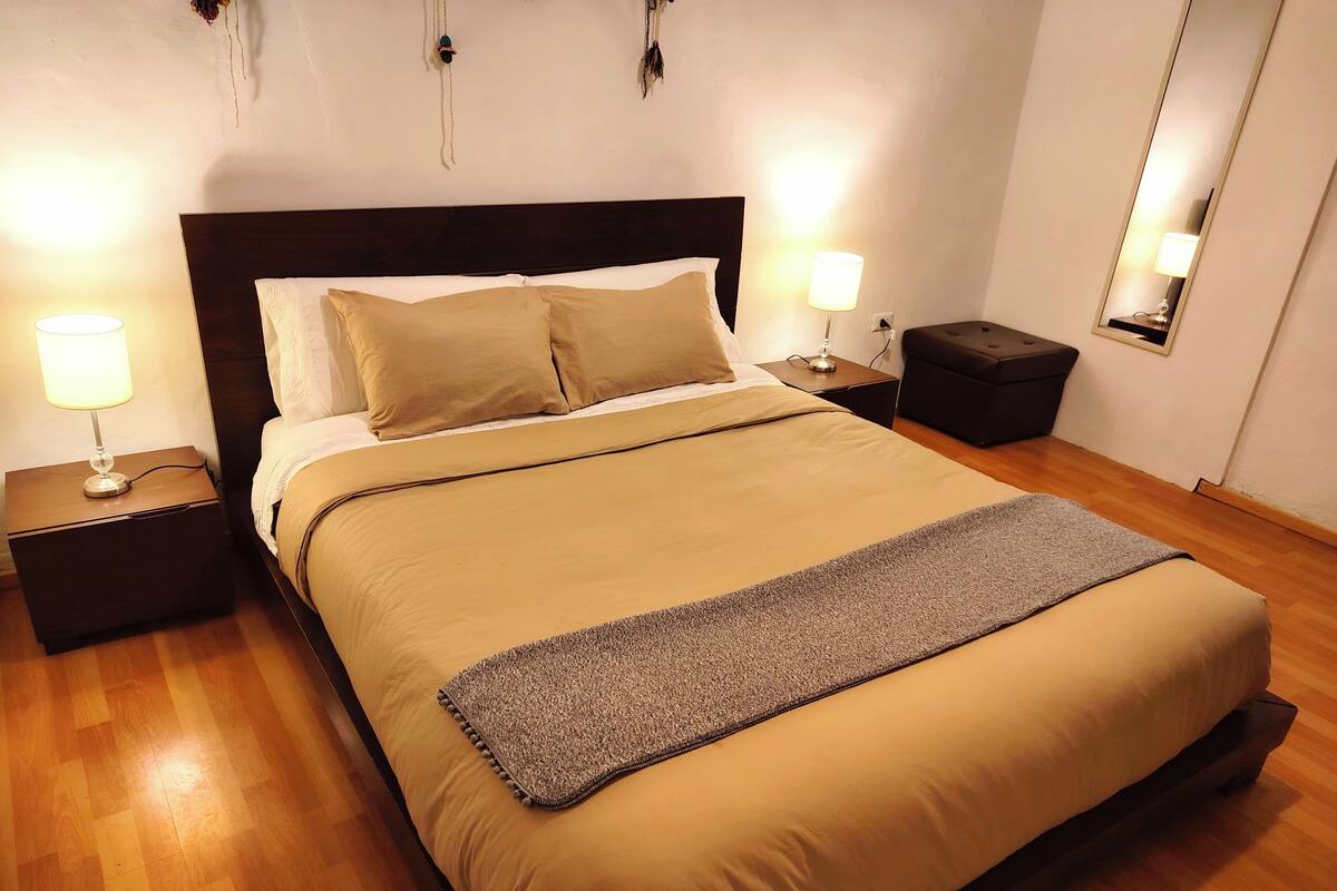 A neatly made bed with a headboard, two bedside tables with lamps, a small nightstand, and a mirror on the wall.