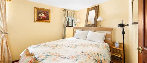 A hotel room with a bed, a bedside table, a lamp, a mirror, and a window with curtains.