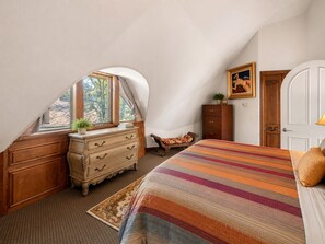 A bedroom with a sloped ceiling, a bed with a striped bedspread, a dresser, a chair, and a painting on the wall. - The Dome (Big Bear City)