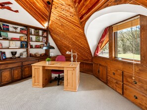 A cozy attic room with a wooden desk, bookshelves, and a large window. - The Dome (Big Bear City)