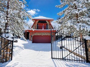 A snow-covered building with a red garage door, surrounded by snow-laden trees and a metal gate. - The Dome (Big Bear City)