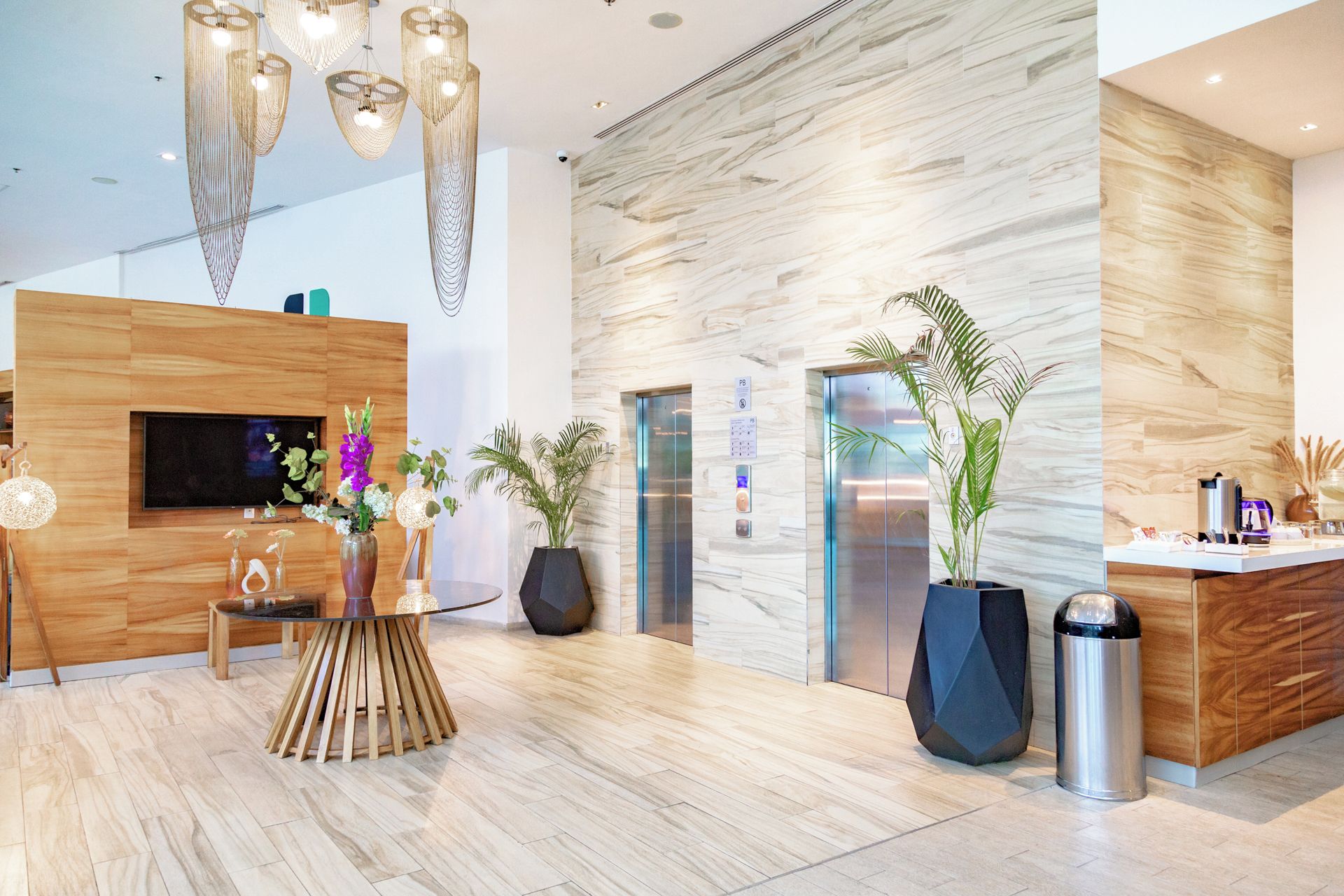A modern lobby with a wooden reception desk, a flat-screen TV, a round wooden table with vases, and potted plants.