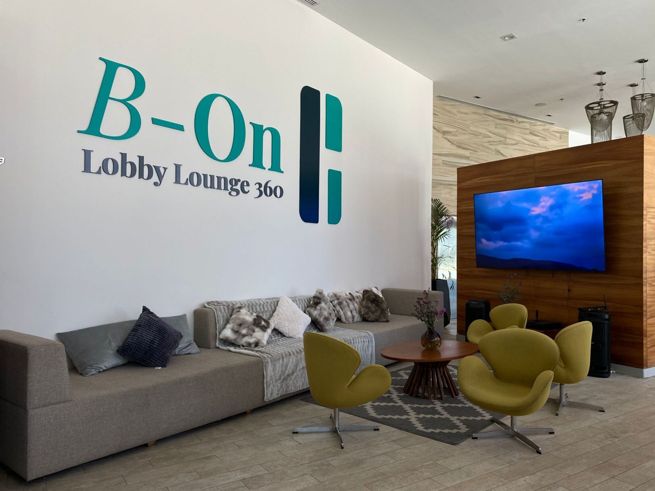 A lobby lounge with a large wall sign reading 'B-On Lobby Lounge 360', a sofa with cushions, two yellow chairs, a round wooden table, and a mounted TV displaying a blue sky.