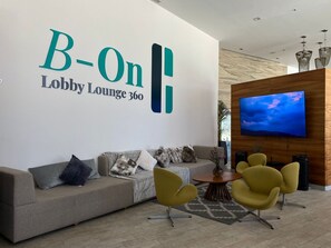 A lobby lounge with a large wall sign reading 'B-On Lobby Lounge 360', a sofa with cushions, two yellow chairs, a round wooden table, and a mounted TV displaying a blue sky. - InHouse Ciudad del Carmen  (Ciudad del Carmen)