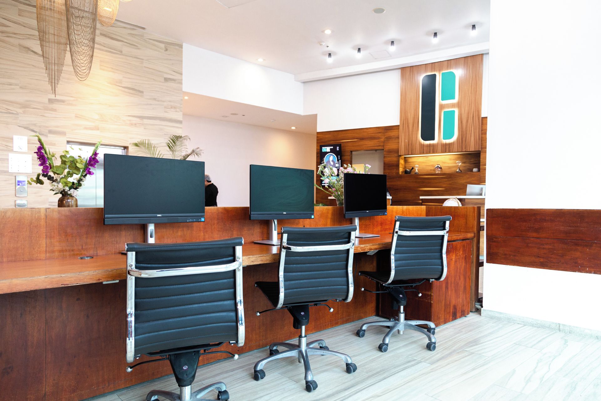 Zona de recepción con escritorio de madera, tres sillas de oficina negras, dos monitores de pantalla plana y un mueble de madera con detalles en color turquesa.