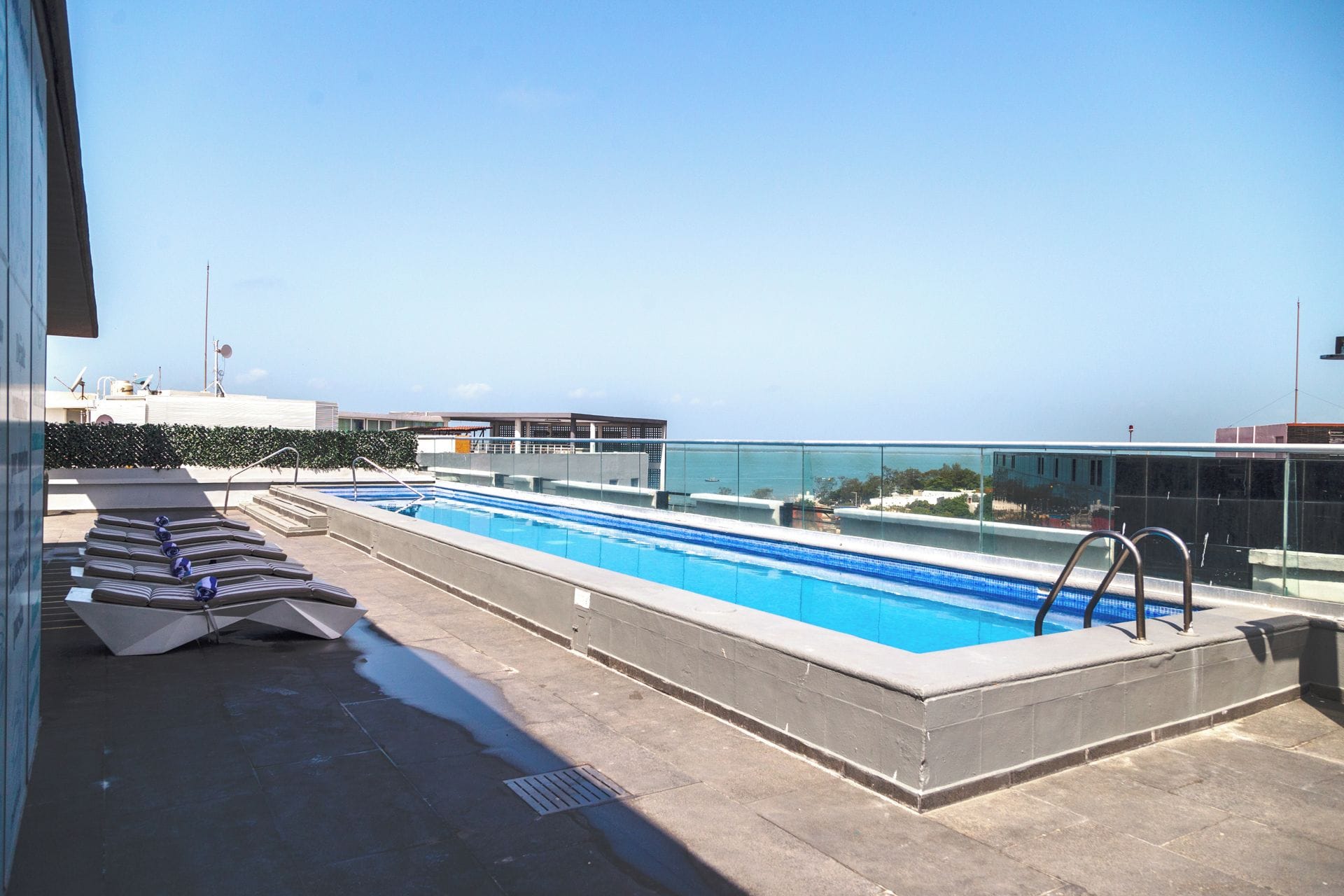 A rooftop swimming pool with a clear view of the sea, a glass barrier, and a set of stairs leading to the pool.
