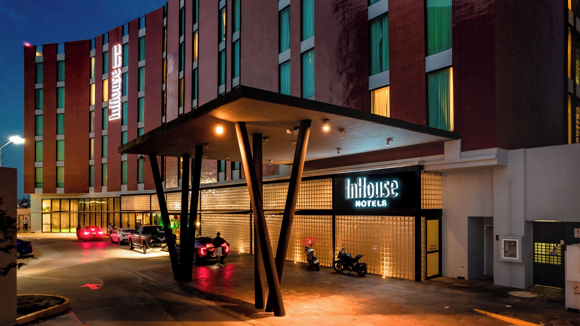 L’entrée d’un hôtel, avec un passage couvert, une enseigne indiquant « Indhouse Hotels » et des véhicules garés.