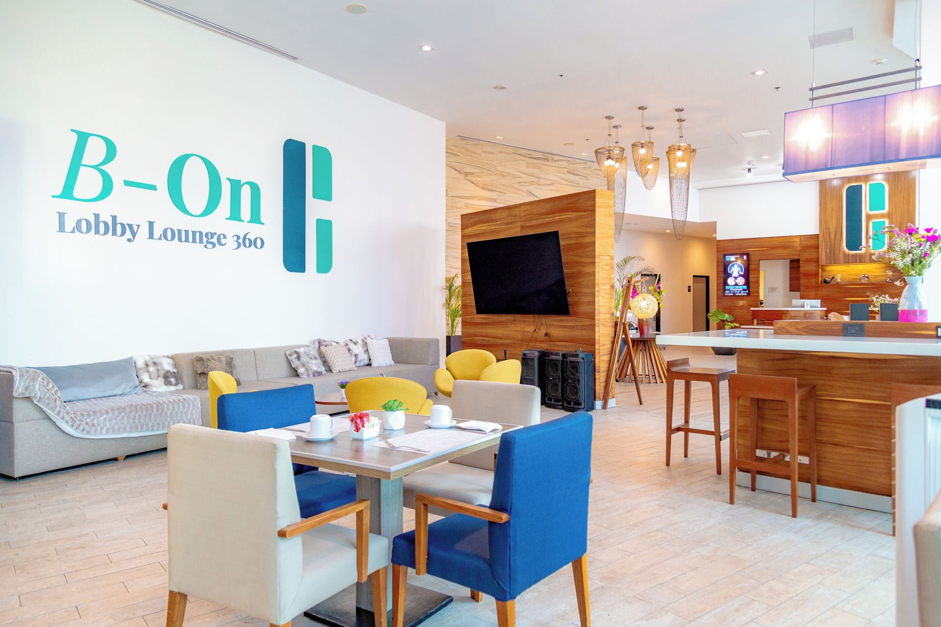 A modern lobby lounge with a sofa, chairs, a table, a TV, and a bar area.