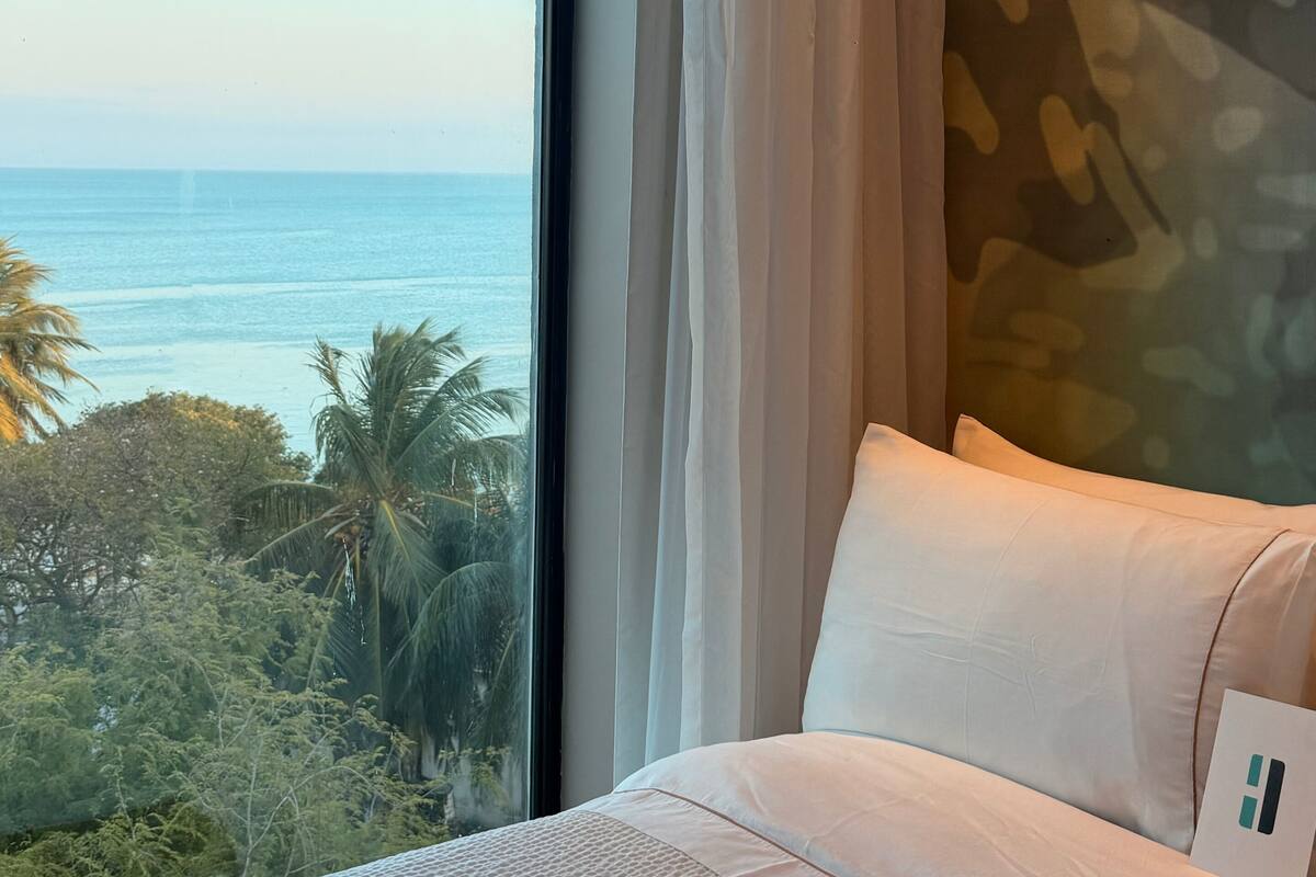 A bedroom with a large window, a bed with white bedding, and a view of the ocean and palm trees.
