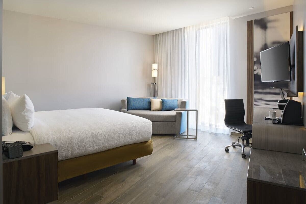 A hotel room with a large bed, a desk, a chair, a TV, and a lamp.