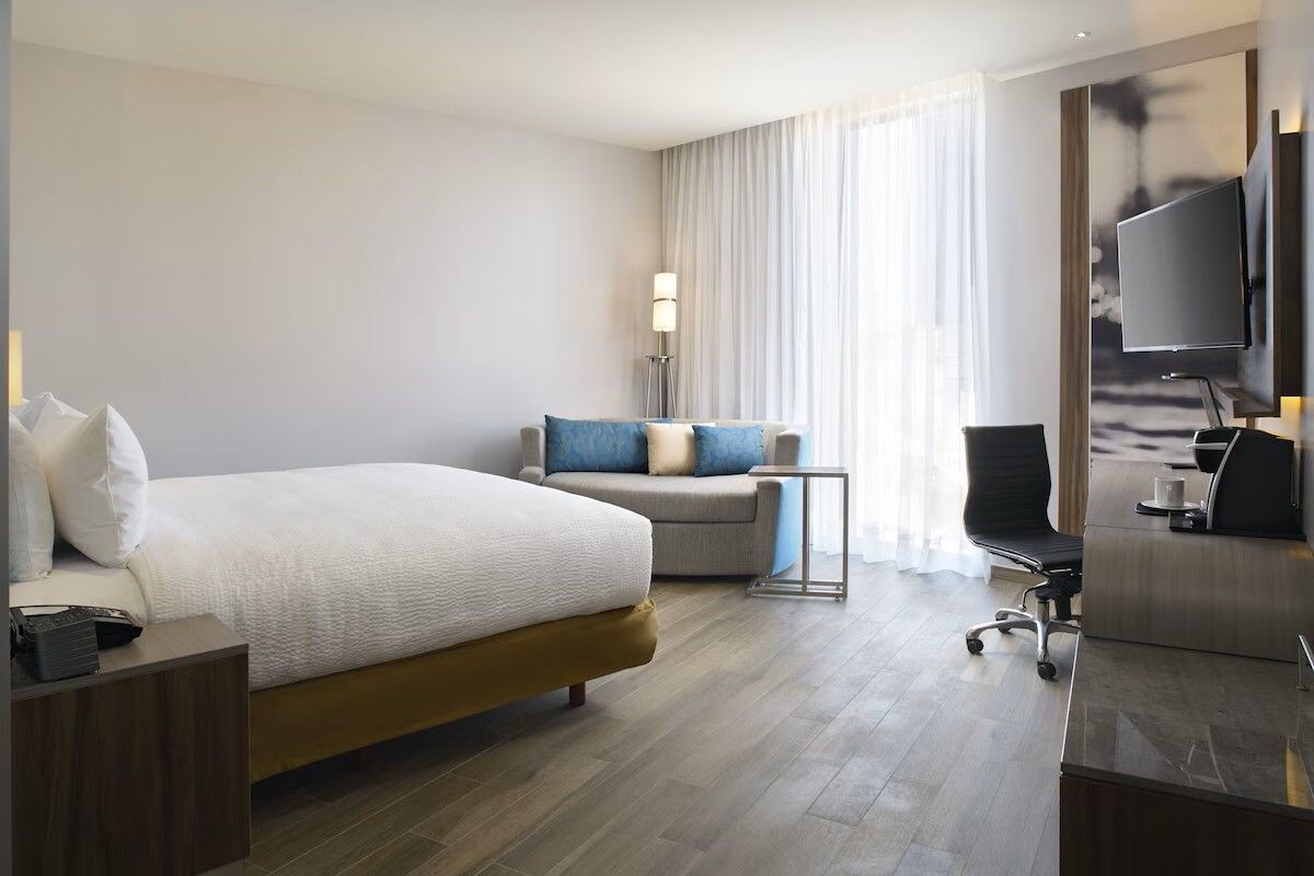 A hotel room with a large bed, a desk, a chair, a TV, and a lamp.