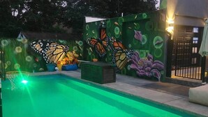 Una piscina con superficie de agua iluminada en verde, rodeada por un muro con un mural de mariposas y flores.