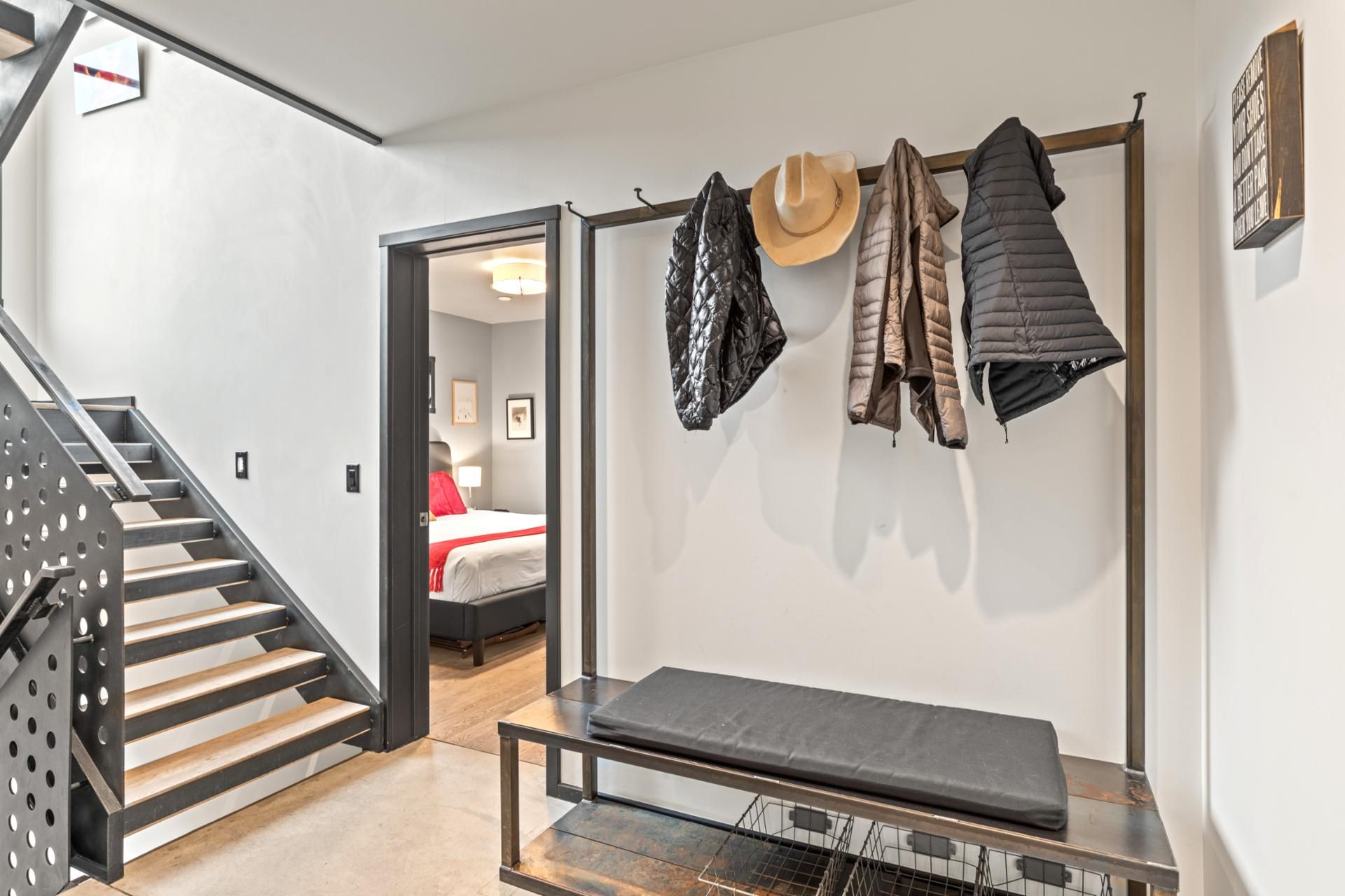 Une chambre d’hôtel moderne avec un escalier, un banc et un porte-manteau sur lequel sont accrochés divers vestes et un chapeau.