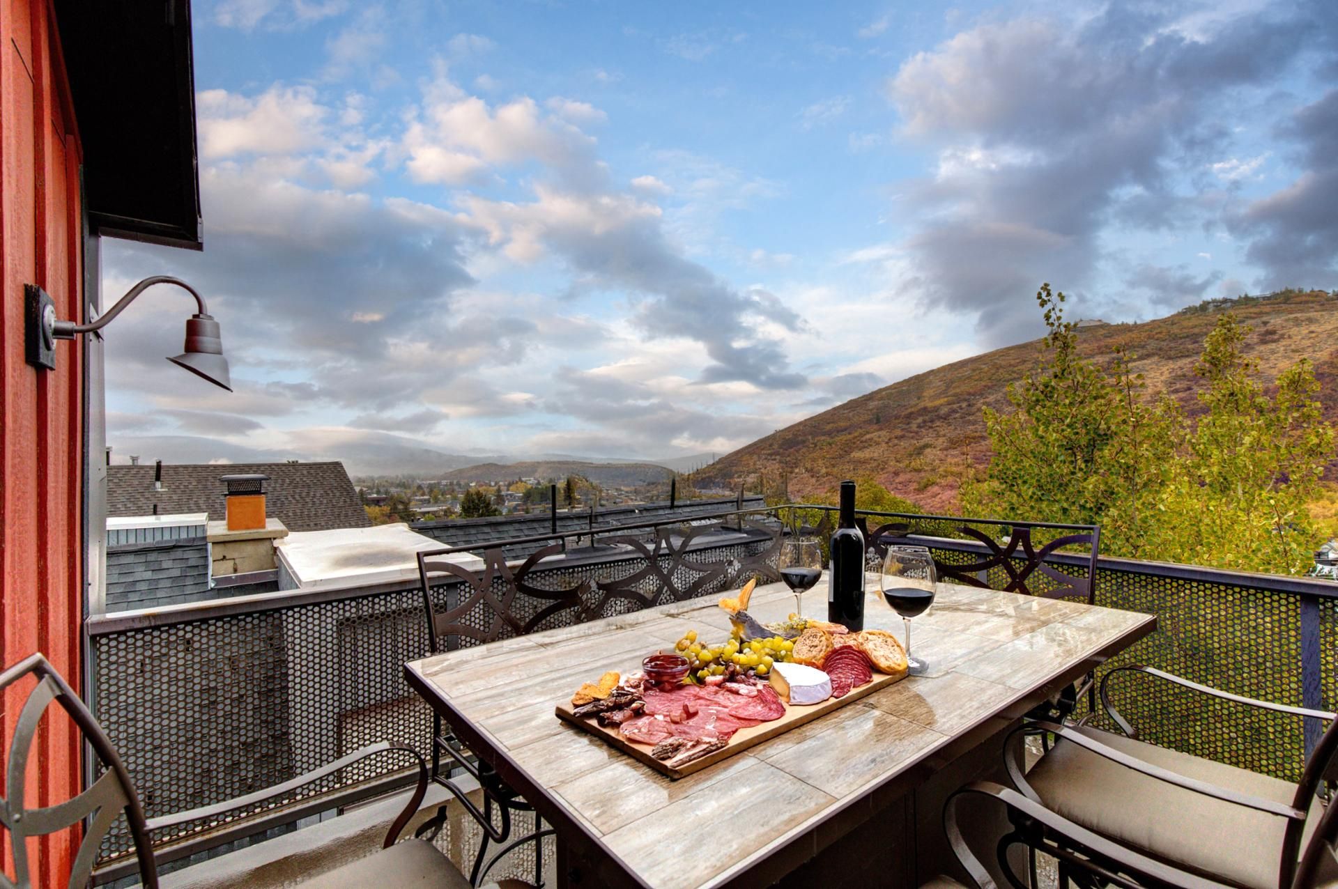 Eine Terrasse mit Tisch, gedeckt für eine Mahlzeit, mit einer Auswahl an Käse, Fleisch und Brot, mit Blick auf eine reizvolle Landschaft mit Hügeln und bewölktem Himmel.
